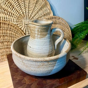 May include: A handmade ceramic pitcher and bowl set. The pitcher has a curved handle and a spout. The bowl is wide and shallow. Both pieces are off-white with brown accents. The set is displayed on a wooden surface.