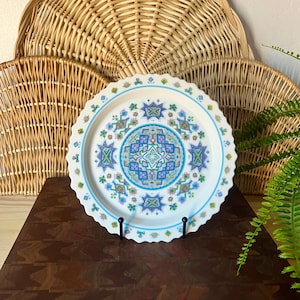 May include: A white decorative plate with a scalloped edge, featuring a geometric design in shades of blue, green, and purple. The plate is supported by a black metal stand, with wicker baskets in the background.