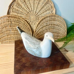 Puede incluir: Una escultura de gaviota de cerámica con cuerpo blanco, alas grises y marrones y pico beige. El ave está posada sobre una tabla de cortar de madera marrón oscuro estampada. En el fondo hay cestas de mimbre y vegetación.