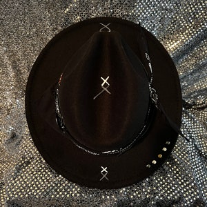 Könnte beinhalten: Schwarzer Filzhut im Fedora-Stil mit einem schwarz-weißen Band. Der Hut hat ein schwarz-weißes X, das auf die Krone und den Hutrand gestickt ist. Der Hut hat ein schwarz-weißes Band mit dem Wort "FRIDAY" darauf.