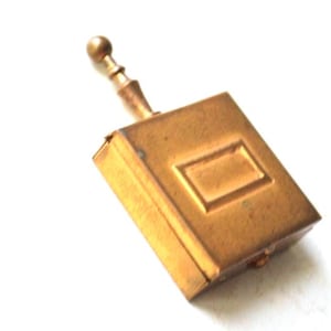 May include: A rectangular brass matchbox holder with a metallic gold finish. The object features a central raised rectangular panel on its front surface and a slender, vertical handle extending from the top, topped with a small spherical finial.