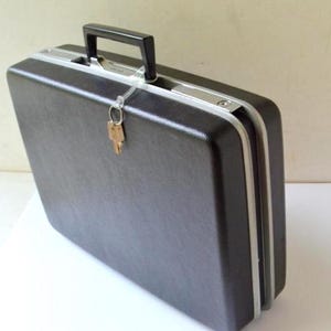 May include: A rectangular black hard-shell briefcase featuring a textured exterior, a black plastic top handle, and a silver-toned metal frame. A single metallic key is attached to the handle with a clear plastic loop, and the case is closed.