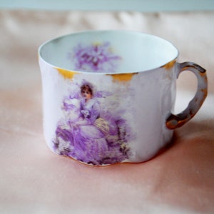 May include: A white porcelain teacup with a gold rim and a floral design featuring a woman in a purple dress. The design is in a vintage style.