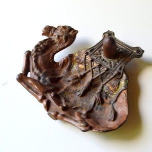 May include: A brown metal tray shaped like a camel with a detailed saddle featuring tassels and a central upright post. The object has a dark bronze-toned finish with gold-colored accents on the saddle area and a textured, sculpted surface.