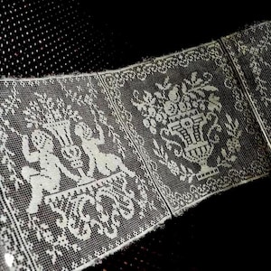 May include: A strip of off-white lace trim featuring a repeating pattern of two figures flanking a central urn filled with flowers. The intricate mesh design includes decorative scrollwork at the base of each panel, creating a continuous border.