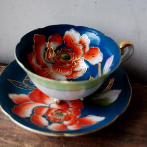 May include: A vintage teacup and saucer set. The cup and saucer are a deep blue with a floral design in red, orange, and white. The cup has a gold handle and trim. The set is likely porcelain.