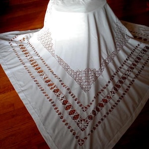 May include: A white, rectangular tablecloth with intricate embroidered patterns. The design features geometric shapes and floral motifs in a contrasting brown color, creating a vintage aesthetic. The tablecloth is draped over an unseen object.