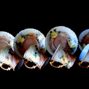 May include: A set of four antique teacups and saucers. The cups and saucers are white with a floral design in yellow, blue, and brown. The cups have gold handles. The saucers have a scalloped edge.