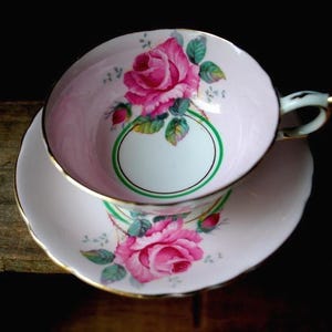 May include: A pink teacup and saucer set with a floral rose design. The teacup and saucer are decorated with pink roses, green leaves, and gold trim. The teacup has a white interior with a green and white oval design. The set is likely made of porcelain.