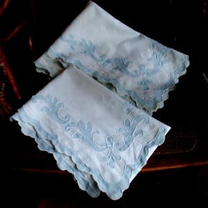 May include: Two white cotton pillowcases with blue embroidered floral designs. The pillowcases have scalloped edges.
