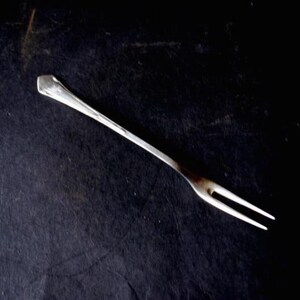 May include: A silver-colored, two-pronged fork with a long, slender handle. The fork is set against a dark, textured background, highlighting its simple design and metallic sheen. Ideal for serving appetizers or desserts.