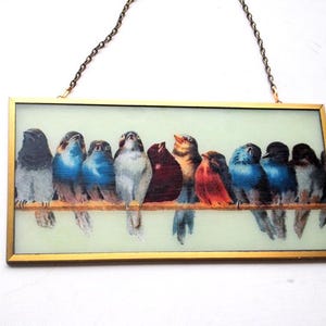 May include: A framed glass panel with a gold frame and a chain for hanging. The panel features a colorful illustration of a row of birds perched on a branch. The birds are a variety of colors, including blue, red, green, and brown.