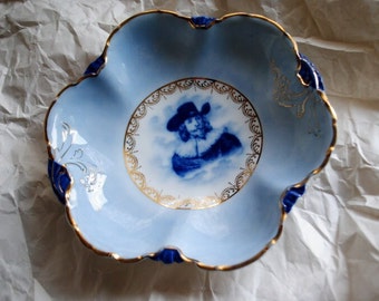 May include: A blue and white porcelain bowl with a gold rim and a central image of a man in a blue and white costume. The bowl has a scalloped edge and is decorated with gold floral designs.