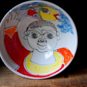 May include: White ceramic bowl with a hand-drawn illustration. The bowl features a smiling face with a yellow collar, red accents, and a fish design. The artwork is outlined in black.