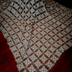 May include: A white and tan lace tablecloth with a geometric pattern. The tablecloth is draped over a round object, showcasing the intricate design. The lace has a diamond and floral pattern, creating a vintage aesthetic.
