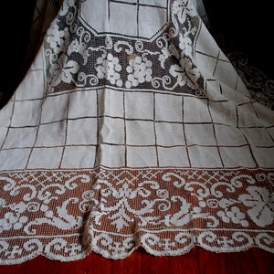 May include: White crocheted tablecloth with a delicate floral pattern and a grid design. The tablecloth has a scalloped edge.
