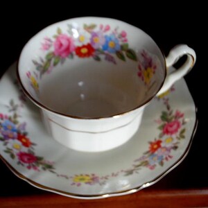 May include: A white teacup and saucer with a floral pattern in shades of pink, yellow, blue, and green. The cup and saucer have a gold trim.