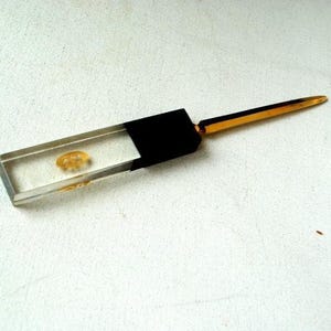 May include: A clear, rectangular letter opener with a small, amber-colored object encased within the handle. The handle transitions to a black section, followed by a gold-colored blade. The opener rests on a white, textured surface.