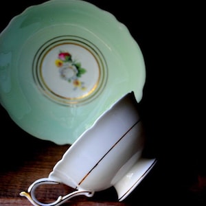 May include: A white teacup with a gold trim and a green saucer with a floral design. The saucer has a scalloped edge.