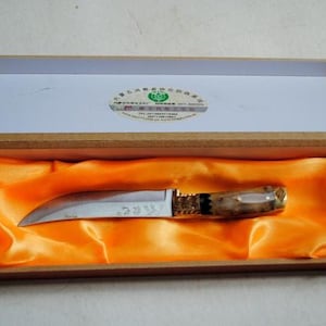 May include: A curved blade knife with a wooden handle and gold accents, presented in a wooden box lined with orange satin. The knife blade has etched text. The box is open, revealing the knife.