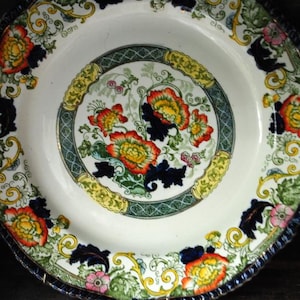 May include: A white ceramic plate with a floral design. The plate features a central floral motif with red, yellow, and orange flowers, surrounded by a green and yellow border. The rim has a dark blue, scalloped edge.