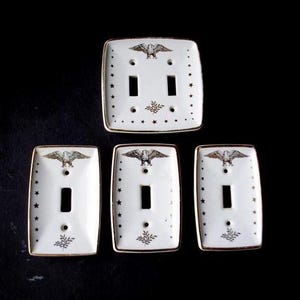 May include: Set of five white ceramic light switch covers. Each cover features a gold-colored eagle, stars, and a decorative leaf design. The edges are trimmed with gold.