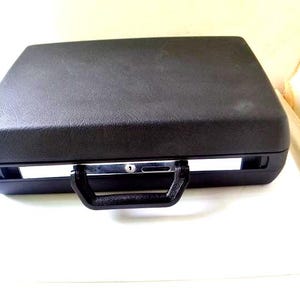May include: A black rectangular hard-shell briefcase with a textured surface. The case features a sturdy black plastic handle attached to the front, a central silver-toned metal locking mechanism, and a slim horizontal silver accent strip.