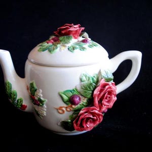 May include: A white ceramic teapot with a lid decorated with 3D roses and leaves. The teapot has a handle and a spout. The roses are pink and red.