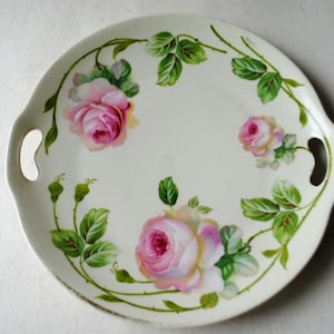 May include: A round, off-white serving tray with two handles. The tray is decorated with pink roses, green leaves, and vines. The floral design is painted around the edge of the tray.