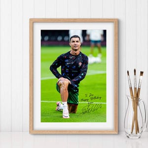 May include: A framed photo of a soccer player wearing a black and green jersey with a colorful pattern. The player is stretching on a green field. The photo is signed "To [Name] Happy Birthday from [Signature]"