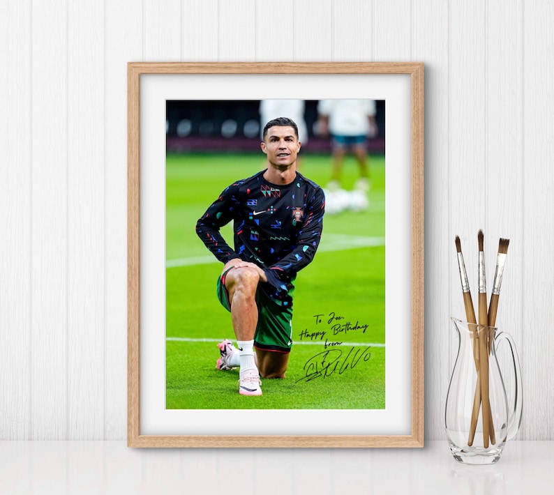 May include: A framed photo of a soccer player wearing a black and green jersey with a colorful pattern. The player is stretching on a green field. The photo is signed "To [Name] Happy Birthday from [Signature]"