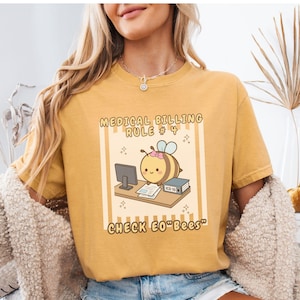 May include: A mustard yellow short-sleeve t-shirt featuring a graphic of a cartoon bee at a desk with a computer and a book labeled ICD-10. The text reads MEDICAL BILLING RULE # 4 and CHECK EO"BEES" in a brown font with a light yellow striped background.