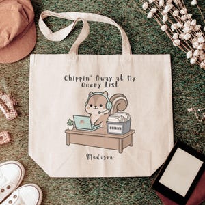 May include: A beige canvas tote bag featuring a cartoon squirrel wearing a headset at a desk with a laptop and a box labelled QUERIES. The text reads Chippin' Away at My Query List and the name Madison. The bag has two top handles.