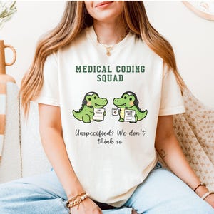 May include: A cream-colored t-shirt featuring the dark green text MEDICAL CODING SQUAD above a graphic of two green crocodiles wearing headsets. One holds a paper labeled LAB RESULTS, the other a coffee mug and DOCTOR'S NOTES. Text below reads: Unspecified? We don't think so.