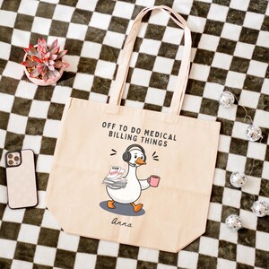 May include: A beige canvas tote bag featuring a cartoon goose wearing a headset, holding a stack of claim papers and a pink mug. The text reads OFF TO DO MEDICAL BILLING THINGS and the name Anna is printed below the goose in black script.