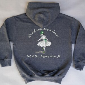 May include: A dark heather gray hooded sweatshirt featuring a graphic of a dancer in a white dress and green hat. The text reads: "it's not easy veing a dancer but if the clogging shoes fit". The design is centered on the back of the garment.