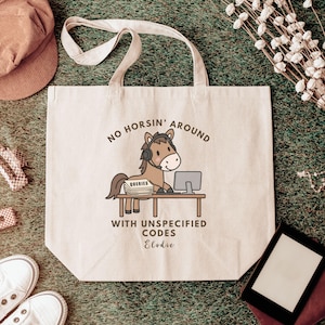 May include: A beige canvas tote bag featuring a brown cartoon horse wearing a headset, sitting at a desk with a computer and a stack of papers labelled QUERIES. The text reads NO HORSIN' AROUND WITH UNSPECIFIED CODES and the name Elodie.