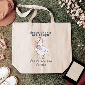 May include: A beige canvas tote bag featuring a graphic of a white duck wearing a pink headband and pink boxing gloves. The text reads, "these charts are tough -but so are you- Caitlin," with small colourful dots surrounding the duck illustration.