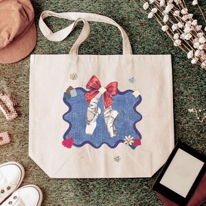 May include: A cream tote bag with a blue denim-style patch displaying ballet pointe shoes and a red bow. The bag has long handles and is decorated with small floral and patchwork accents. The bag is on a green surface.