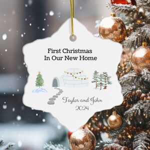 May include: White snowflake-shaped Christmas ornament with a white igloo, two pine trees, and a string of colorful lights. The text reads "First Christmas In Our New Home" and "Taylor and John 2024".