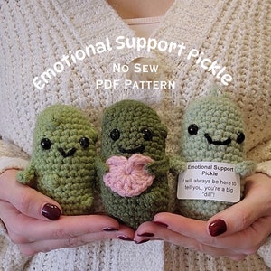 May include: Three crocheted pickle plushies, one holding a pink heart. The plushies have smiling faces and black eyes. The text on the tag reads "Emotional Support Pickle I will always be here to tell you, you're a big "dill"!"