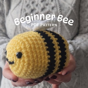 Puede incluir: Un juguete de abeja de crochet con rayas negras y amarillas, alas blancas y ojos negros. La abeja está siendo sostenida en las manos de alguien. El texto "Beginner Bee PDF Pattern" está por encima de la abeja.
