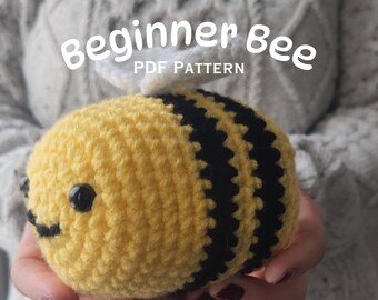 Puede incluir: Un juguete de abeja de crochet con rayas negras y amarillas, alas blancas y ojos negros. La abeja está siendo sostenida en las manos de alguien. El texto "Beginner Bee PDF Pattern" está por encima de la abeja.