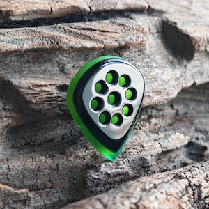May include: A guitar pick with a green, black, and silver design. The pick has a teardrop shape with a silver center featuring seven green circular accents. The edges are green, and the pick is set against a textured wooden background.