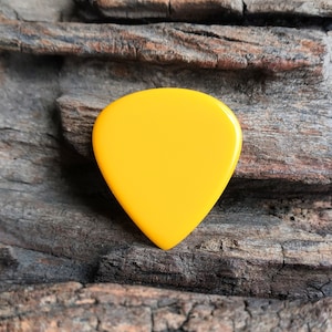 Può includere: Un plettro per chitarra di colore giallo brillante, realizzato in un materiale liscio e lucido. Il plettro ha la forma di una lacrima e si trova su un pezzo di legno ruvido e marrone.