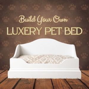 Puede incluir: Cama de perro de madera blanca con un cojín estampado en blanco y marrón. La cama tiene un borde elevado y un ribete decorativo. El texto "Build Your Own LUXURY PET BED" está escrito encima de la cama.