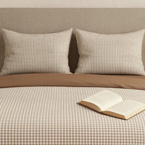 Könnte beinhalten: Ein Bett mit neutralem Farbschema, mit einem Kopfteil, zwei Kissen und einem Bettbezug, alles in einem hellbraunen und weißen Vichy-Muster. Zwei passende Lampen mit Holzsockeln stehen auf Nachttischen. Ein offenes Buch liegt auf dem Bett.