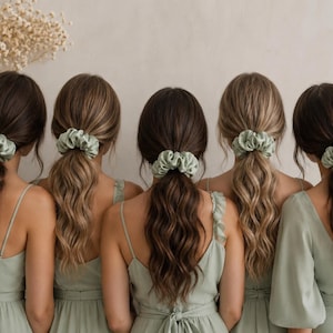 May include: Five individuals seen from behind, each wearing a sage green satin scrunchie in their hair. The hair is styled in low ponytails with loose waves. Each person is wearing a matching sage green dress with varying sleeve styles.