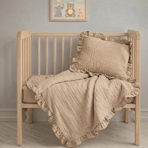 Könnte beinhalten: Ein hellbeiges Musselin-Bettwäscheset bestehend aus einem rechteckigen Kissen und einer Decke, beide mit Rüschenkanten. Die Bettwäsche liegt in einem hellen Holzbettchen, darüber befindet sich ein gerahmtes Bild mit Elefant, Bär und Fuchs.