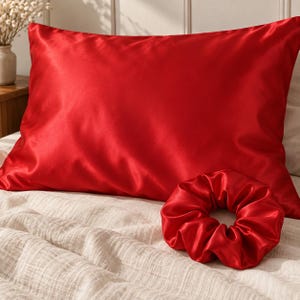 May include: A rectangular pillowcase in a vibrant red colour with a smooth, lustrous finish, accompanied by a matching red hair scrunchie. Both items feature a consistent, solid red tone and a soft, reflective texture, presented together on a bed.